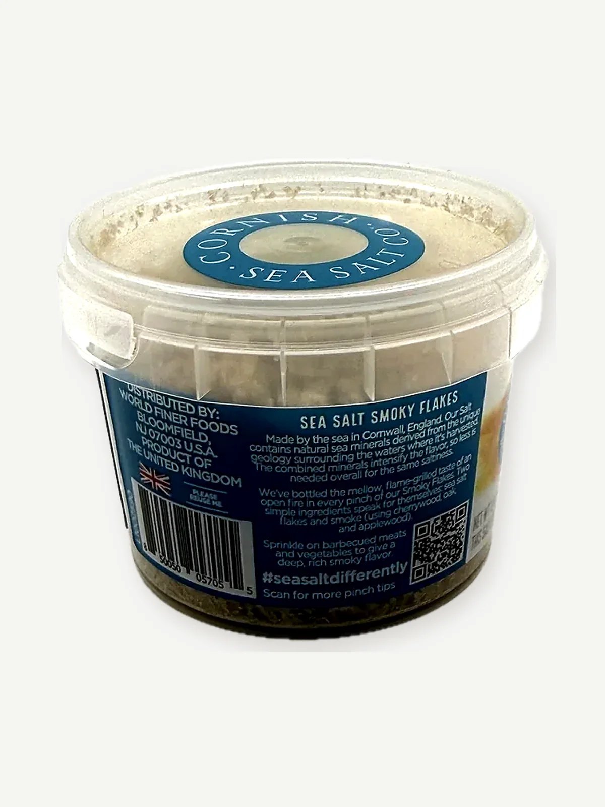 Back of a clear plastic tub of Cornish Sea Salt Co. Smoky Flakes showing the label story panel, distributor details and barcode
