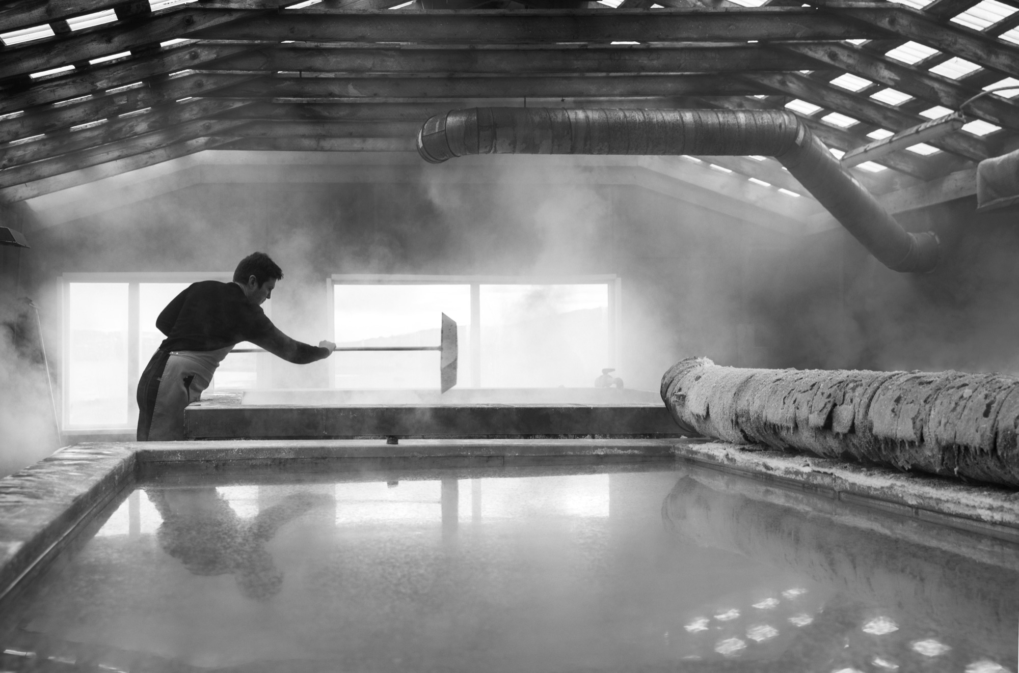 Black-and-white photograph of a Saltverk saltmaker working over steaming pans at Reykjanes