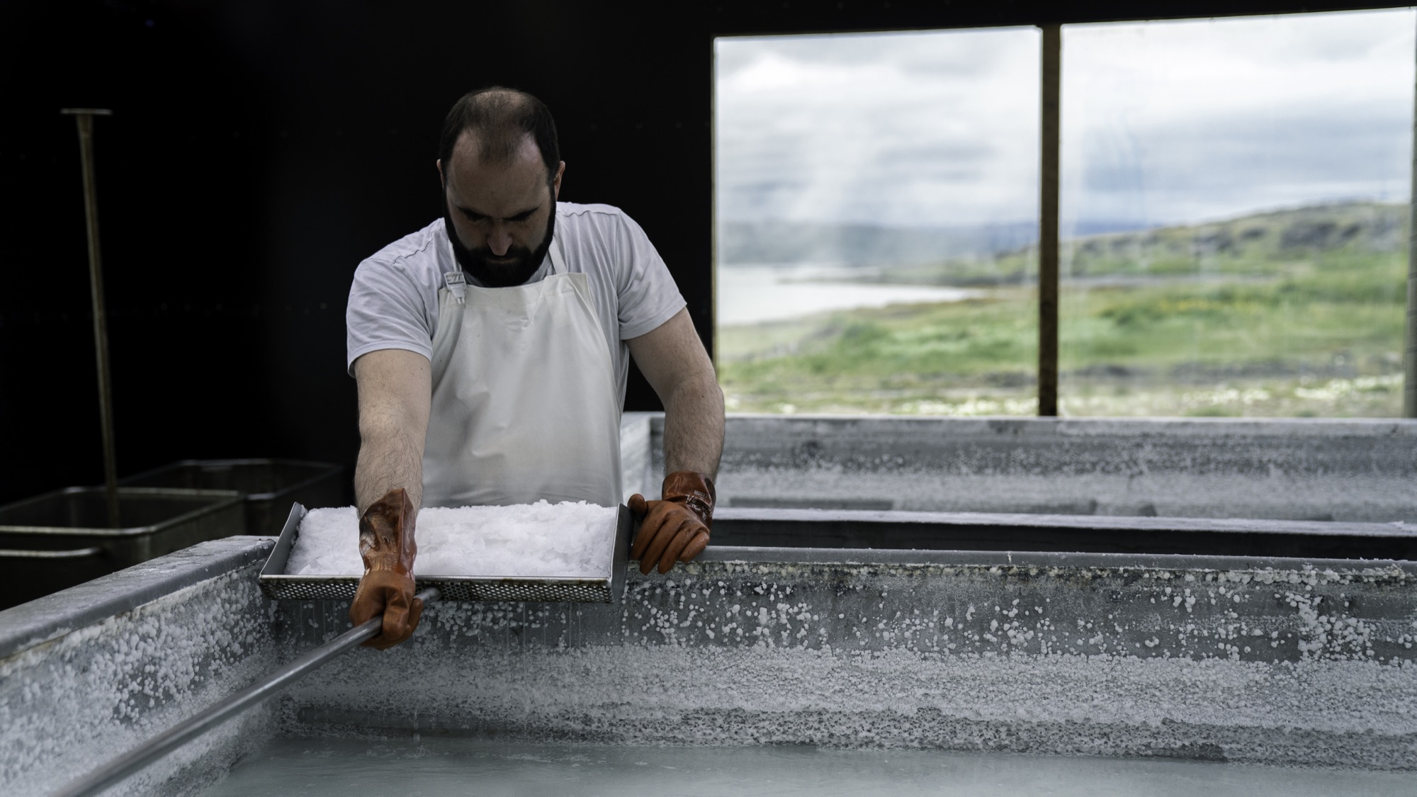 A saltmaker in a white apron lifts a tray of wet salt crystals from a crusted salt pan, with a window onto the fjord behind him