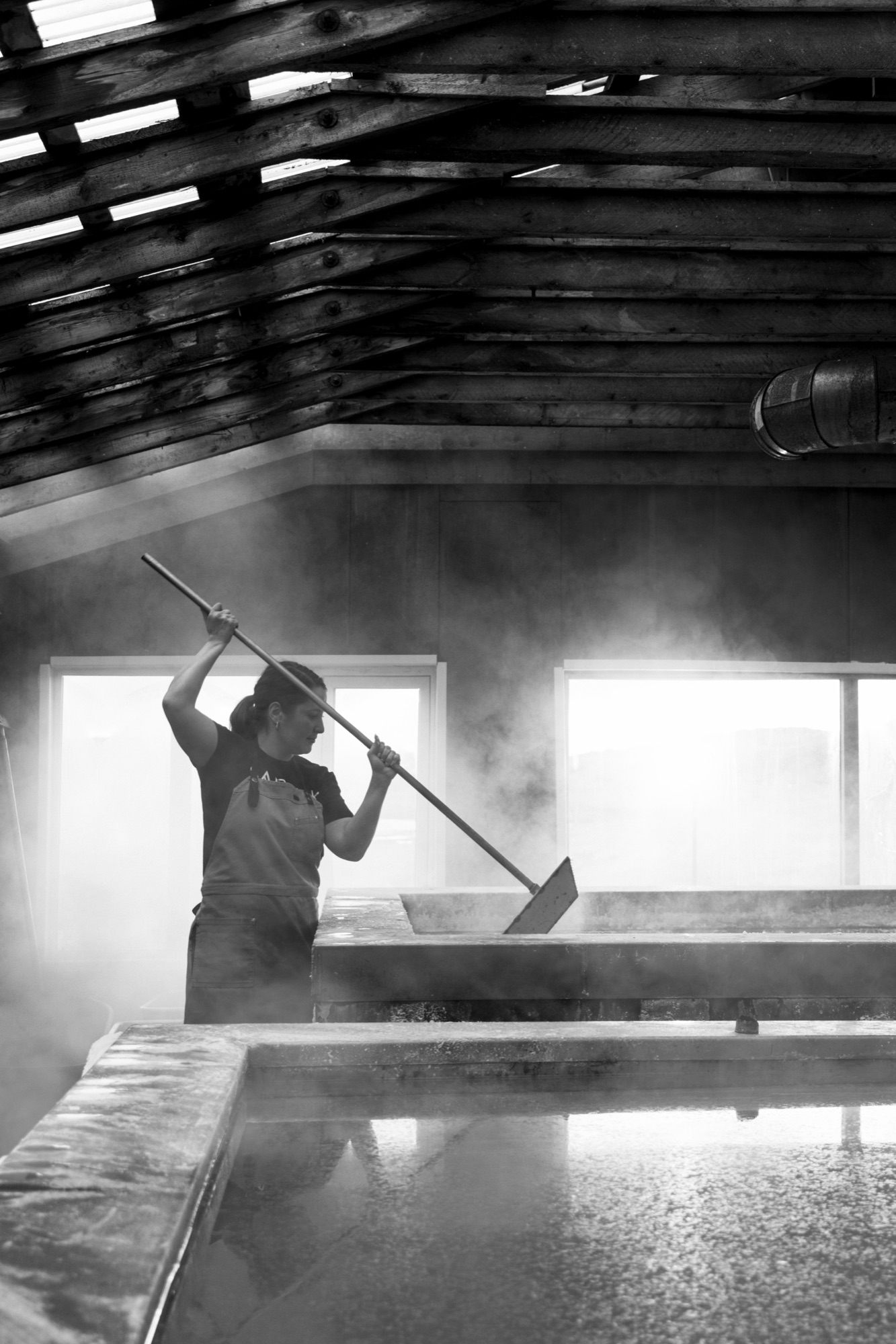 Black-and-white portrait of a Saltverk saltmaker using a long-handled rake over a steaming pan